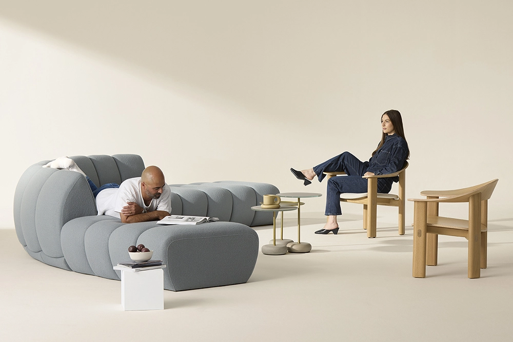 soft seating in a sparse, but well-lit room. a man lies on the sofa reading a magazine, while a woman sits in a curved wooden lounge chair looking at the nest of coffee tables in front of her.